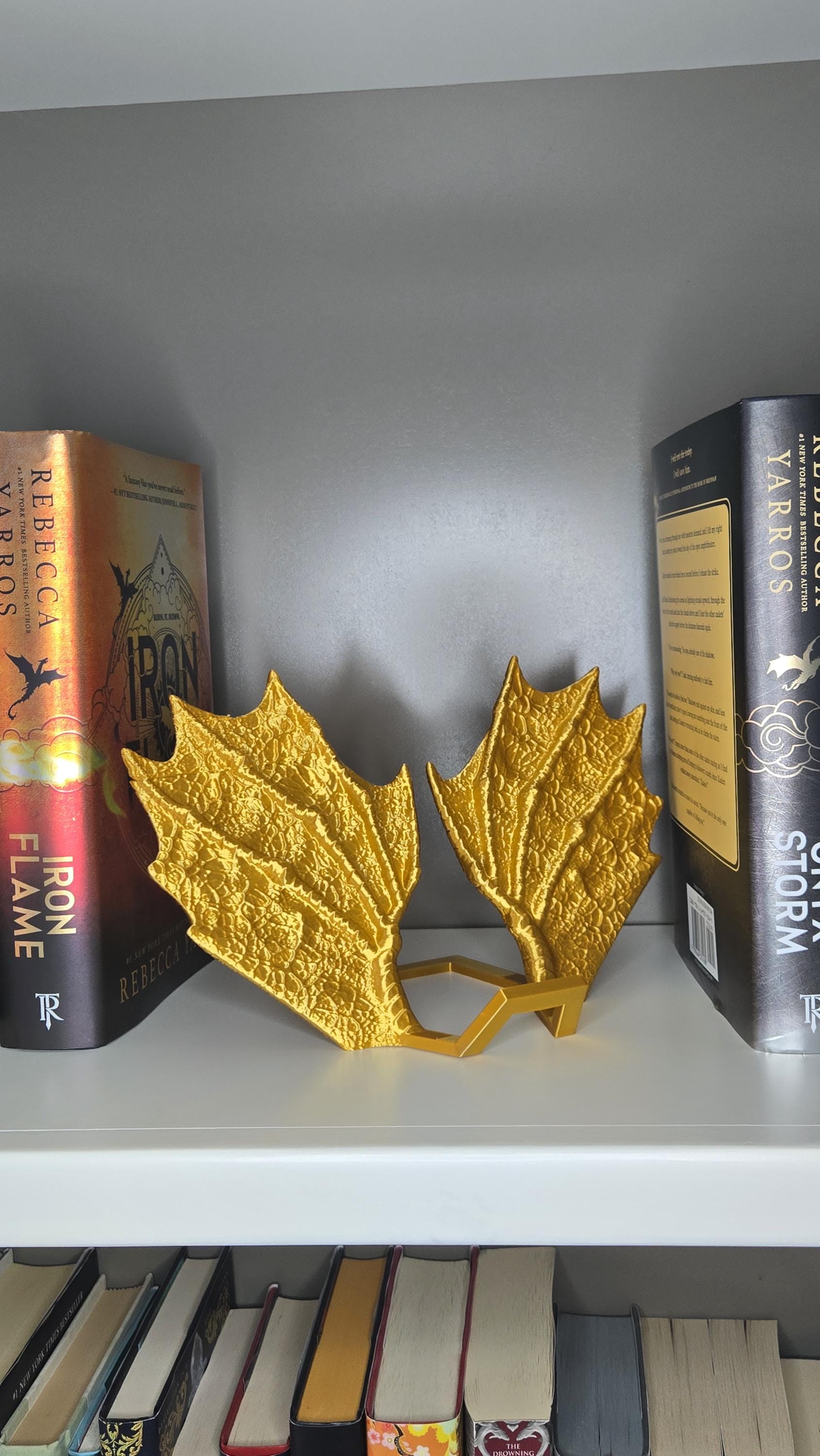 May include: Two gold dragon wings with a textured surface. The wings are displayed on a shelf with two books on either side. The books are titled 'Iron Flame' and 'Storm'.